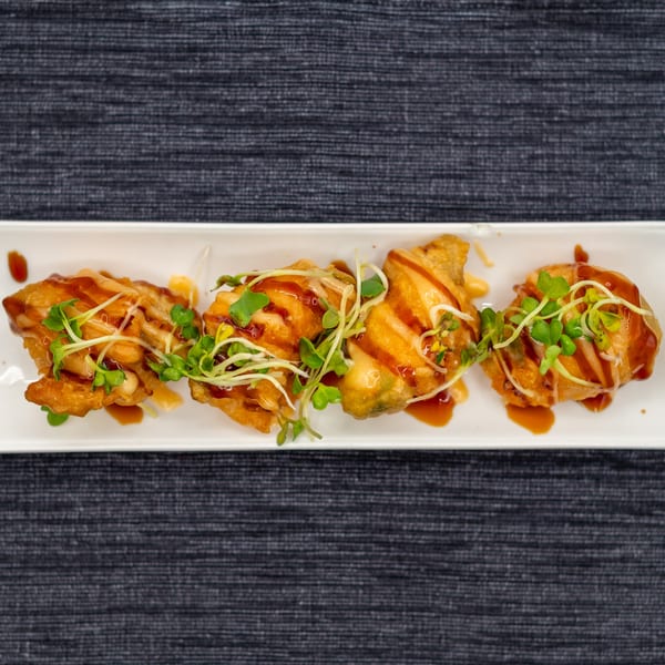Four pieces of fried sushi, drizzled with sauce and topped with microgreens, on a white plate.