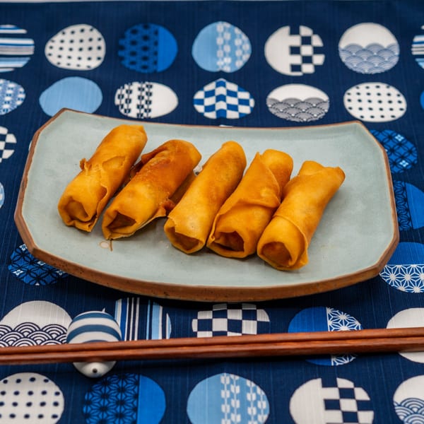 Five crispy golden spring rolls on a light blue plate with chopsticks on a blue patterned tablecloth.