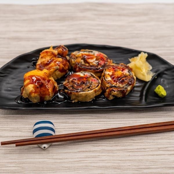 Fried sushi rolls drizzled with sauce, served on a black plate with ginger, wasabi, and chopsticks.