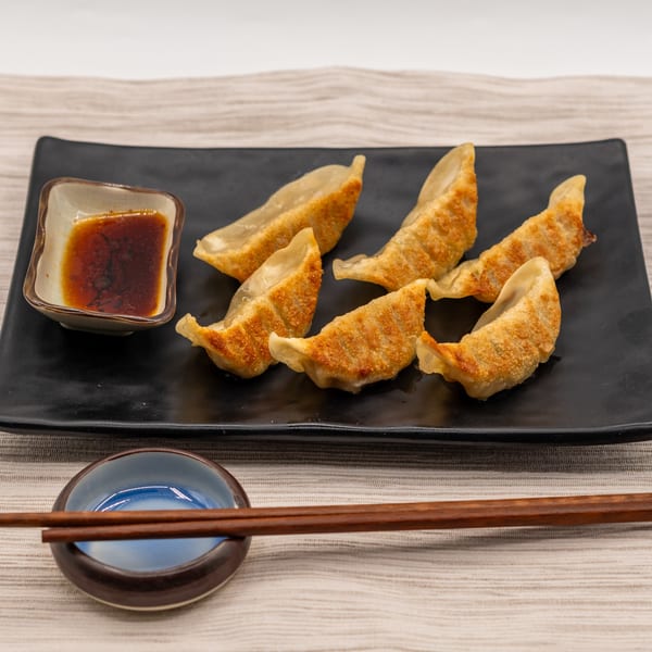 Six golden-brown gyoza dumplings with a side of dipping sauce and chopsticks on a black plate.