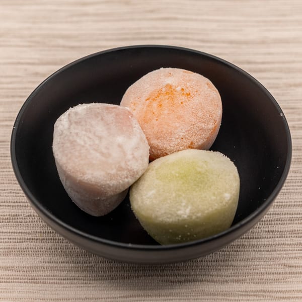 Three colorful mochi ice cream balls, orange, pink, and green, in a black bowl.