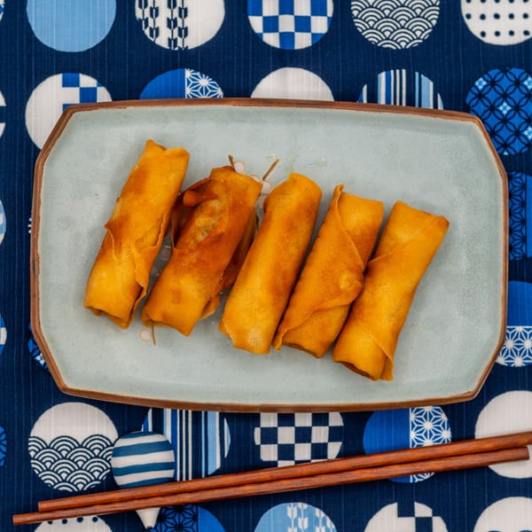 Five golden spring rolls on a light blue rectangular plate with chopsticks on a blue patterned tablecloth.