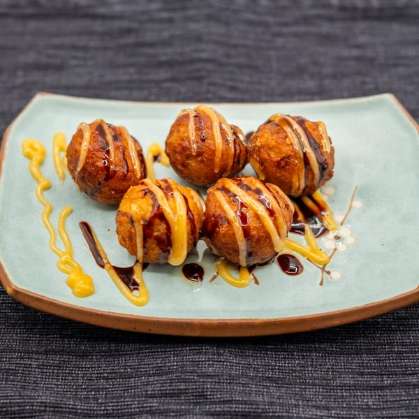 Five golden-brown takoyaki balls drizzled with dark and light sauces on a light blue plate.