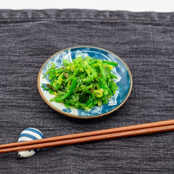 A small blue and white bowl of bright green seaweed salad with sesame seeds, next to chopsticks.