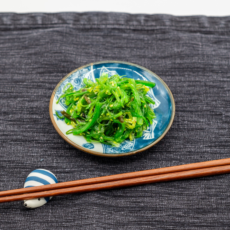 A small blue and white bowl of bright green seaweed salad with sesame seeds, next to chopsticks.